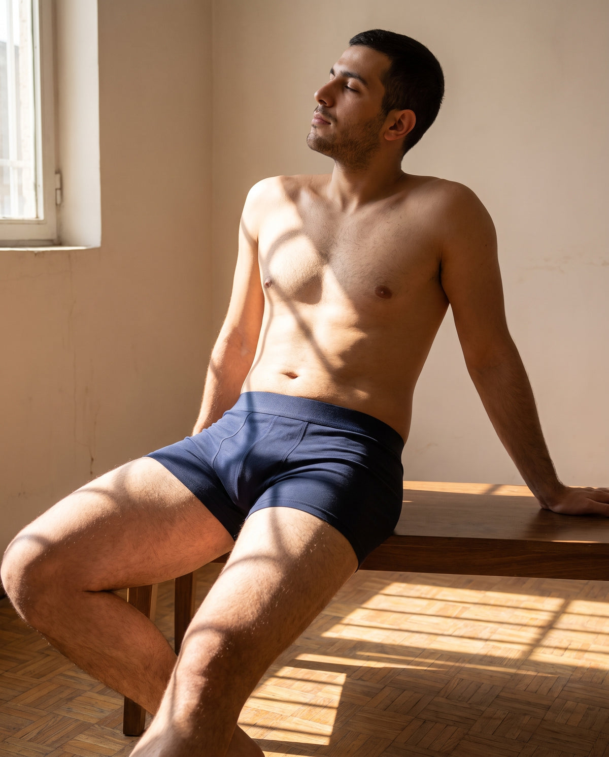 A man seated on a bench in hard afternoon sun, wearing the KAYA navy cotton trunk
