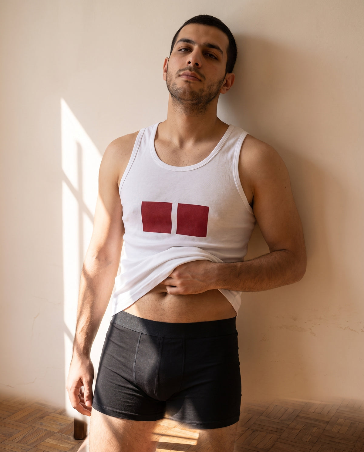A man against a sunlit plaster wall in the KAYA white printed cotton undershirt and black cotton trunk